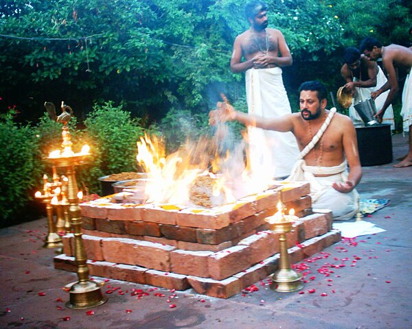 Indian man sitting in front of a rectangular brick altar with a burning fire. The altar is surrounded by candles as he throws a powder into the flames.